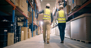 Two workers wearing high-visibility vests and hard hats walk through a warehouse aisle lined with shelves of boxes and cartons.