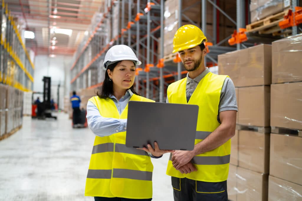 Two warehouse workers wearing safety vests and helmets look at a laptop together in a storage facility with shelves and stacked boxes.