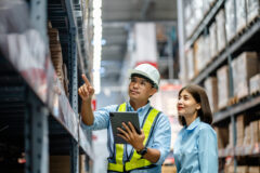 A man in a safety vest and helmet points at shelves in a warehouse while holding a tablet. A woman stands beside him looking in the same direction.