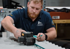 A man wearing a blue shirt operates a tool on a piece of ridged metal in an industrial setting.