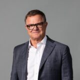A middle-aged man wearing glasses, a white shirt, and a gray plaid suit jacket stands against a plain light gray background, looking at the camera and smiling slightly.