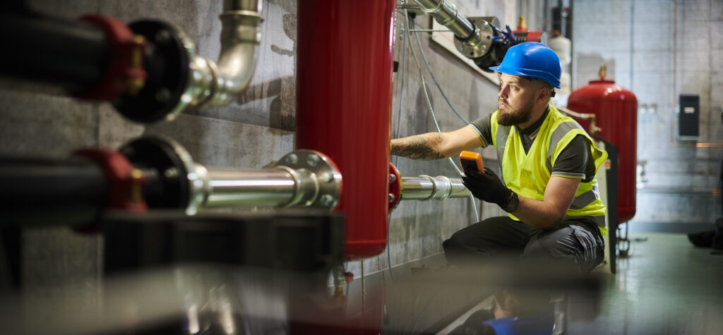 A worker in a safety vest and blue helmet inspects industrial pipes and equipment, using a handheld device, inside a modern industrial facility.