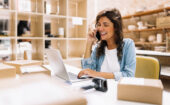 Woman sitting at a desk with a laptop, talking on the phone and smiling, surrounded by cardboard boxes and office supplies.