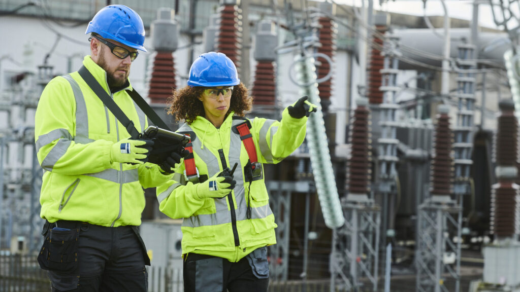 Two utility workers in high-visibility jackets and blue helmets inspect equipment at an outdoor electrical substation.