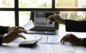 Two people review financial documents at a desk with a laptop displaying charts and graphs, a calculator, and papers spread out in front of them.