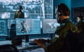 A person in military uniform monitors multiple computer screens displaying maps and data in a command center with large digital displays in the background.