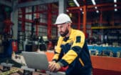 A man in safety gear and a hard hat uses a laptop in an industrial warehouse with machinery and metal structures in the background.