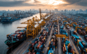 Aerial view of a busy shipping port with numerous colorful containers, large cranes, docked cargo ships, and a city skyline under a cloudy sky at sunset.