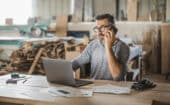 Man with glasses and beard sits at a desk with a laptop, talking on the phone in a woodworking workshop, surrounded by papers and tools.