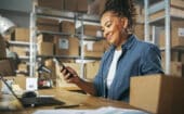 A woman sits at a desk in a warehouse, smiling while looking at her smartphone. Cardboard boxes, shelves, a laptop, and paperwork are visible around her.