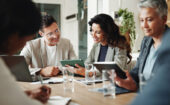 Four people sit at a table with tablets, laptops, and papers, engaged in discussion during a meeting in a modern office setting.