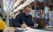 A man sits at a desk in a warehouse, writing in a notebook and using a laptop, with shelves of goods and workers in the background. A yellow hard hat is on the table.