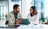 Two people sit at a desk in an office, having a discussion while one holds a tablet. Laptops, papers, and glasses of water are on the table.