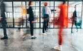 Blurred image of several people walking inside a modern office with glass walls and visible furniture in the background.