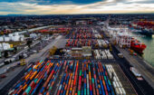 Aerial view of a busy shipping port with numerous colorful shipping containers, cranes, and industrial buildings under a partly cloudy sky.