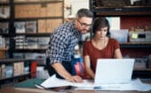 Two people in casual clothing review documents and work together on a laptop in a warehouse or office with shelves and boxes in the background.