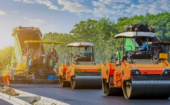 Construction workers operate heavy machinery to pave and smooth a new road, with trees and blue sky in the background.