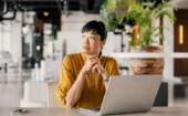 Person in a yellow shirt sits at a desk with a laptop, holding a pen and looking thoughtfully to the side in a modern office setting.