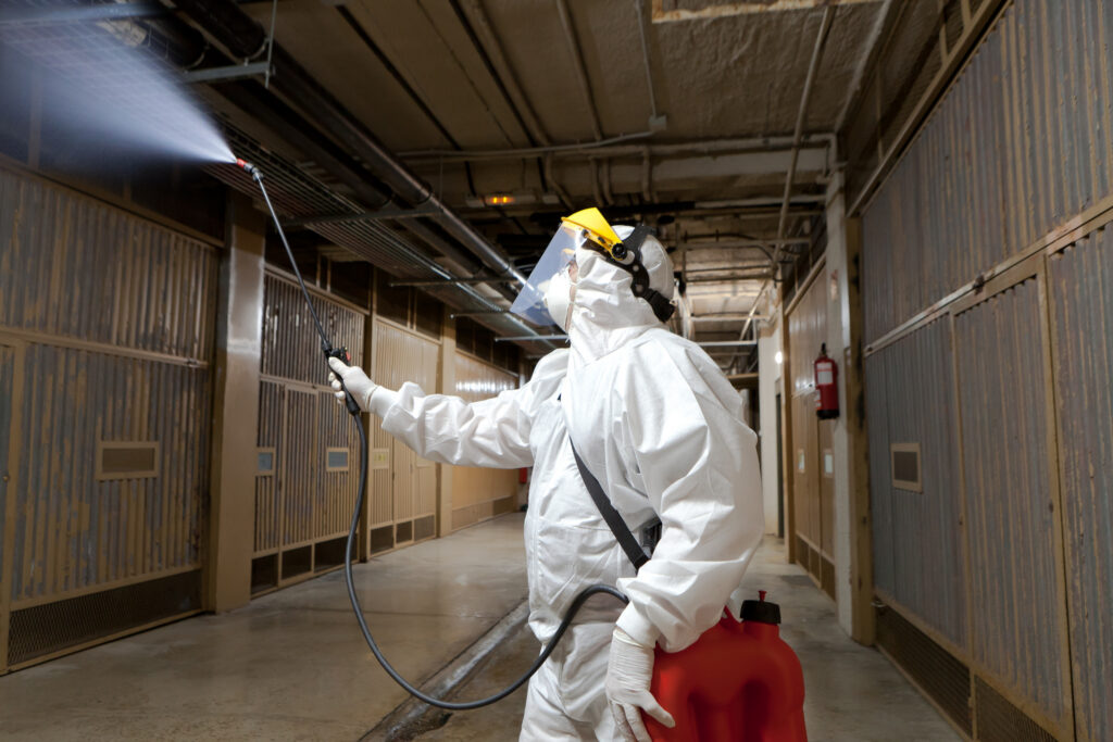 A person in protective gear sprays disinfectant on the ceiling of an industrial hallway or facility.