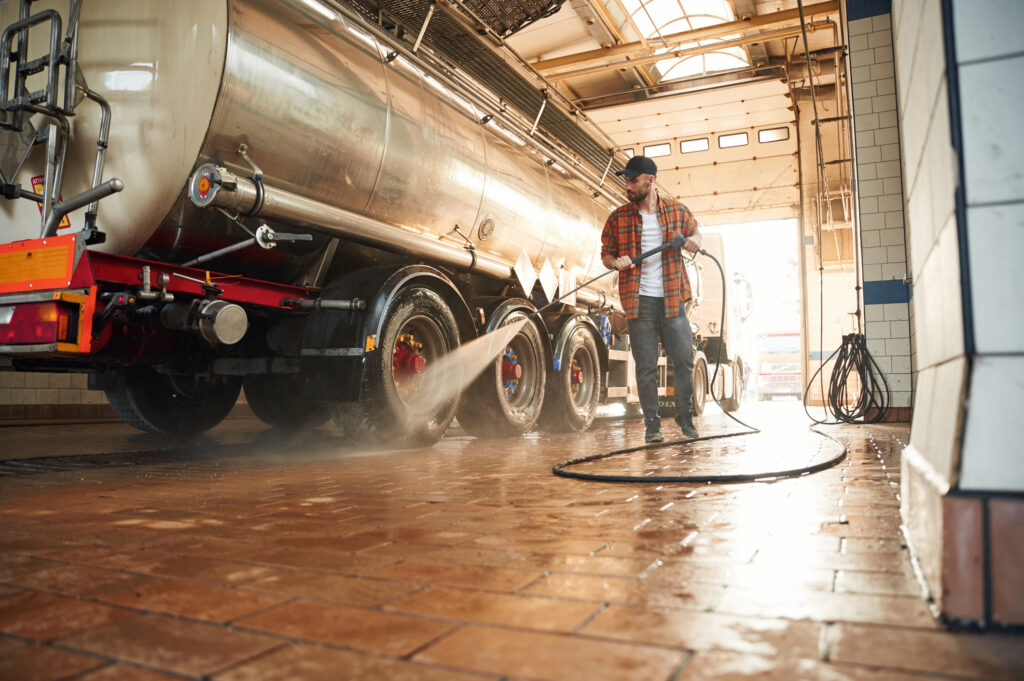 A man uses a pressure washer to clean the tiled floor next to a large tanker truck inside a garage or washing facility.