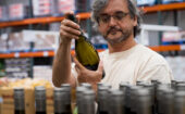 A man in glasses examines a bottle of wine in a warehouse aisle, with shelves of products in the background.