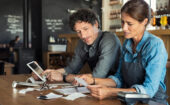 Two people in aprons sit at a table reviewing receipts and papers, with a tablet and payment terminal nearby in a cafe or restaurant setting.