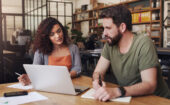 Two people sit at a desk in an office, discussing work while looking at a laptop. One person types, and the other takes notes with a pen and notepad.