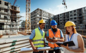 Three construction workers in safety gear review documents and blueprints at an active building site with cranes and unfinished structures in the background.