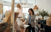 Woman standing in a store, talking on the phone while using a laptop at a wooden counter surrounded by shelves and wicker baskets.