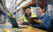 Two warehouse workers sit at a desk with a laptop and clipboard, reviewing documents. Shelves of inventory and other workers are visible in the background.