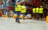 Warehouse workers in yellow safety vests walk and move among pallets and shelves stocked with various goods and materials.