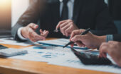 Three people in business attire review financial charts and graphs at a desk, using pens, a calculator, and documents during a meeting.