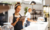 Woman in apron uses a tablet in a bright café kitchen, while another person in the background arranges items near a glass display with pastries.