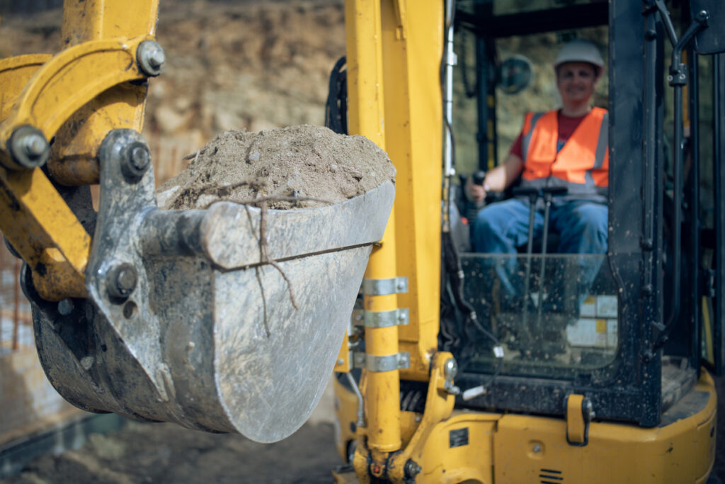 A construction worker operating a yellow excavator lifts a bucket of dirt at an outdoor worksite.