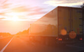 A yellow semi-truck drives on a highway at sunset, with sun glare and lens flares visible in the image.