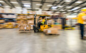 Worker operates a yellow forklift transporting boxes inside a warehouse, with shelves of packages and another worker in the background.