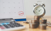 A calendar with “Deadline” circled on the 30th, a stack of coins, a calculator, and an alarm clock on a wooden desk.