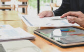 Two people in business attire review printed charts and graphs at a desk with a tablet, papers, and office supplies.
