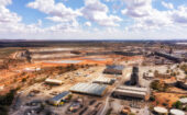 Aerial view of a large mining site with industrial buildings, open pits, machinery, and reddish tailings ponds surrounded by arid landscape under a partly cloudy sky.
