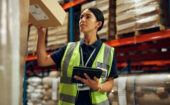 A woman in a safety vest holds a box and checks a tablet in a warehouse with shelves of packages in the background.