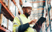 A man in a high-visibility jacket and hard hat uses a tablet in a warehouse with shelves of boxes in the background.