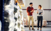 Two people walk and talk in a warehouse, surrounded by stacked pallets and shelves. One holds a tablet, suggesting a discussion or inventory check.