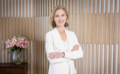 A woman in a white suit stands with arms crossed, smiling, in front of a wood-paneled wall. A vase of pink lilies sits on a table behind her.