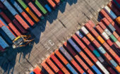 Aerial view of colorful shipping containers arranged in rows at a cargo port, with a crane visible on the left side.