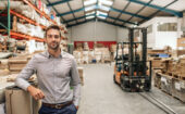 Man in business attire stands in a warehouse near stacked shelves and a forklift, looking at the camera.