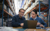 Two people sit at a desk with a laptop in a warehouse, discussing work. Shelves filled with boxes and supplies are visible in the background.