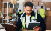Man wearing a safety vest checks a tablet and documents at a warehouse workstation, with other workers in the background handling boxes and shelves.