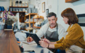 Two people wearing aprons sit at a counter, reviewing information on a tablet, with shelves and various items visible in the background.
