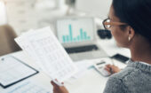 Person wearing glasses reviews printed financial documents at a desk with a laptop displaying bar charts, calculator, and other papers.