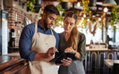 A man and a woman in aprons stand behind a bar, looking at a tablet together.